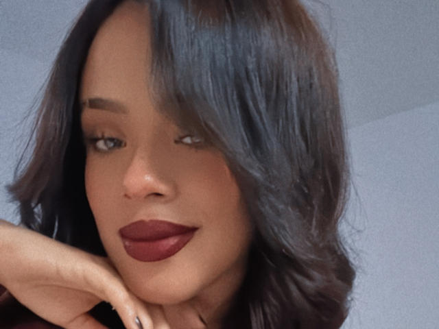 Lia Hilton a les cheveux longs, bruns et ondulés qui lui cachent un œil, elle porte un rouge à lèvres rouge vif et a un sourire charmant. Elle repose son menton sur sa main, et elle a l'air séduisante et confiante..
