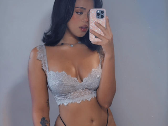 LiaHilton with dark hair, wearing a lace bra that shows her boobs, black thong, holding a pink phone, tattoo on arm, blue necklace, slightly parted lips.