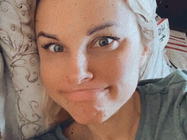BelleLisaG with blonde hair, blue eyes, and a slight smile, lying down, wearing a green shirt, with small dark freckles on her face, looking at the camera.