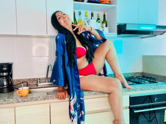 JackieMature with black hair, red bra and panties, blue floral robe, sitting on kitchen counter, leg raised, making a peace sign, smiling, barefoot, bottles in background.