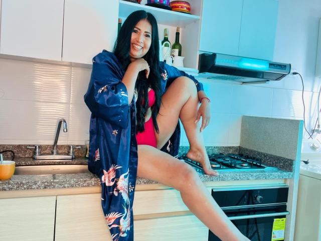 JackieMature with long black hair, smiling, wearing a blue floral robe, pink lingerie, and no pants, sitting on a kitchen counter with one leg up, showing her bare pussy.