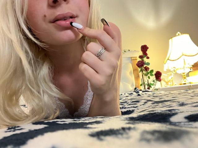 LaPulia with blonde hair, white lace top, and black-white nail polish, biting her finger, showing a ring, lying on a blue-white patterned bed, with a lamp and roses.