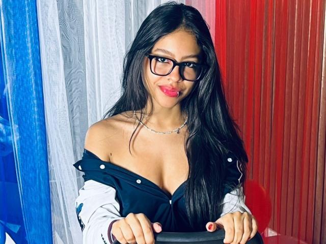 SkyWinter with long black hair, glasses, red lipstick, and a low-cut black top showing her tits. She's wearing a silver necklace and has red nails.