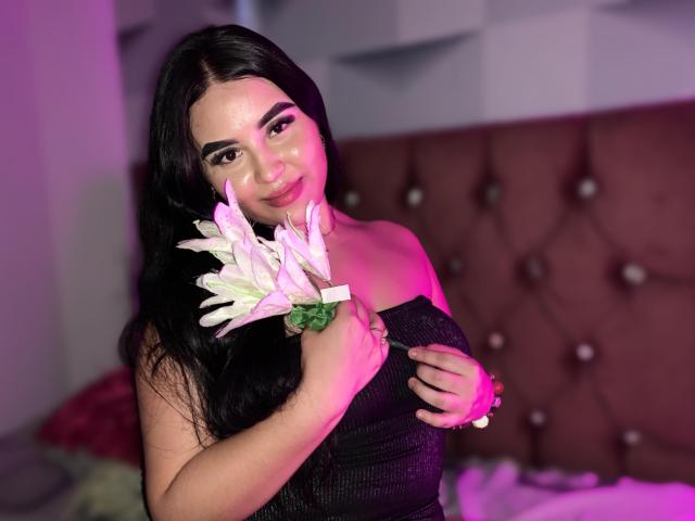 IsabellaaSweett with long black hair, holding white flowers, wearing a black strapless dress, pink and purple lighting, smiling, brown tufted headboard, and pink pillow in the background.