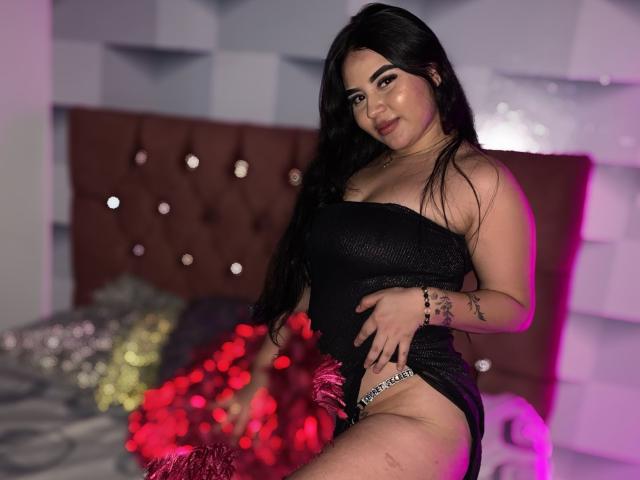IsabellaaSweett, con lunghi capelli neri, indossa un abito nero senza spalline, che lascia intravedere la sua biancheria intima colorata, seduta su un letto con luci rosse, mentre guarda la fotocamera..