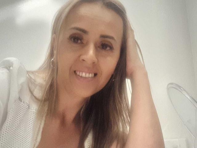 MaylinYung with long blonde hair, smiling, wearing a white top, hand in hair, showing her teeth, earrings visible, in a slightly blurry selfie.