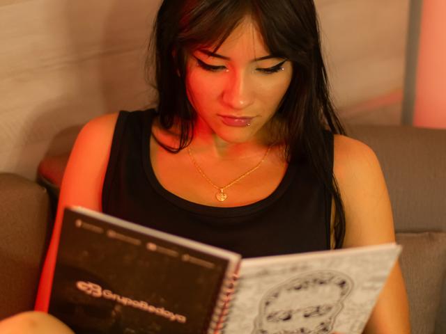 LizzeJoy with dark hair, wearing a black sleeveless top and gold necklace, reading a book with red lighting on her face, showing a focused expression. Her tits are partially visible.