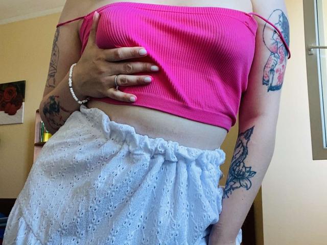 KarKutie's pink ribbed top, showing her small tits, with white lace pants, has tattoos on both arms, a white bracelet, and a hand on her belly.