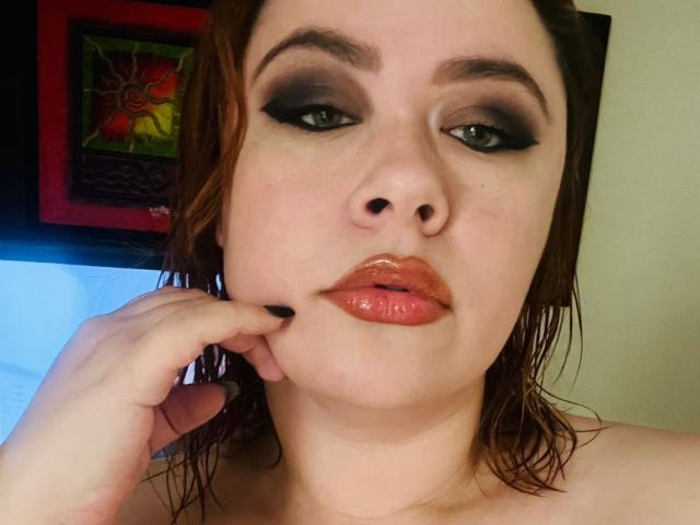 MaddyBlooms has glossy pink lips, dark smoky eye makeup, and wet brown hair, touching her chin with a black-painted nail. Her blue-green eyes are slightly open.