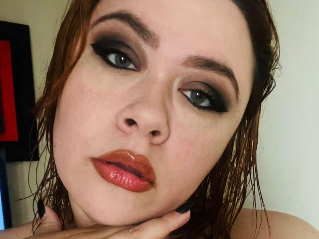 MaddyBlooms has wet brown hair, dark eye makeup, and glossy orange lips. She's looking at the camera with her hand near her chin, showing black nail polish.