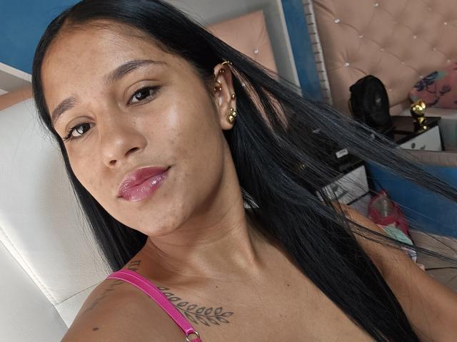 MichelWod has dark brown skin, long black hair, gold earrings, pink lipstick, and a tattoo on the right shoulder. She's wearing a pink strap, looking at the camera.