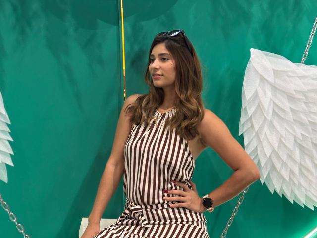 Sara Hills con un vestido a rayas marrón y blanco, cabello castaño ondulado, gafas de sol en la cabeza, alas de ángel blancas, mano en la cadera, fondo verde, usando un reloj de pulsera negro..