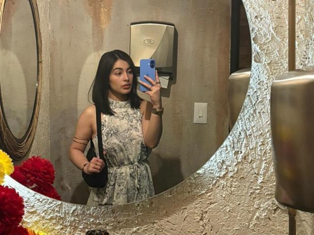 EmmaBeaumont with medium tits, wearing a floral dress, holding a blue phone, taking a mirror selfie, textured mirror, black bag, brown wall, and a paper dispenser in the background.
