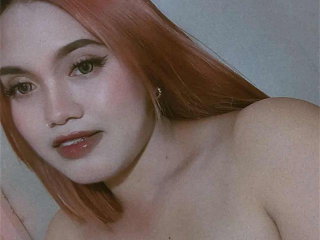 AsianHotCock69 a de longs cheveux roux, des yeux verts, est nue et a de petites seins ronds et fermes, porte un boucle d'oreille en or, et a des lèvres roses légèrement entrouvertes et brillantes..