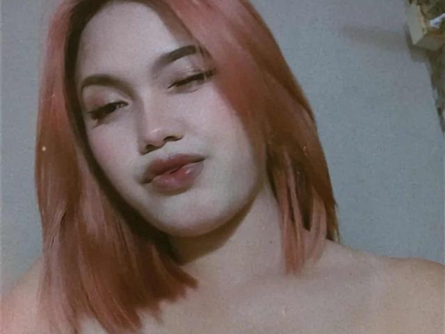 AsianHotCock69 mit rosa Haaren, voller Lippen und heller Haut, blickt leicht lächelnd nach unten und zeigt ihre nackten Schultern. Weiches, körniges Bild, leicht unscharf..