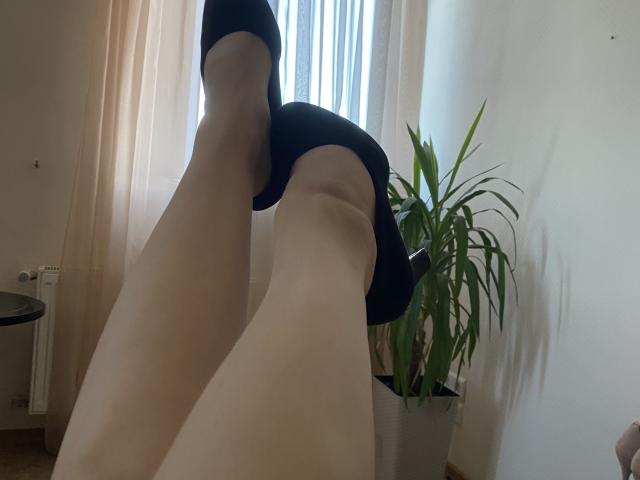 Olieri's pale legs in black heels, legs crossed, showing smooth skin and black shoes, with a green plant to the right and beige curtains in the background.