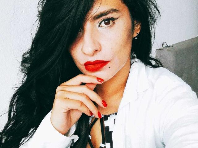 MoserratRouse has long black hair, red lipstick, and red nail polish, wearing a white shirt, with a hand near her face, brown eyes, and a small mole on her cheek.