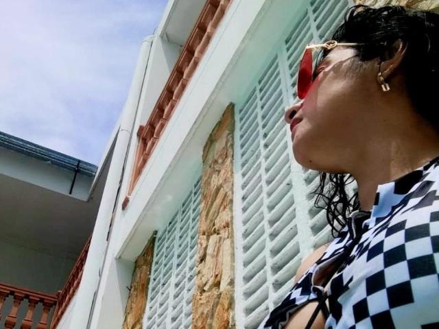 Moserrat, con cabello oscuro y ondulado, gafas de sol rojas y una blusa a cuadros, está de pie cerca de contraventanas blancas y una pared de piedra, mostrando sus pequeños y lindos senos, con un cielo azul soleado..