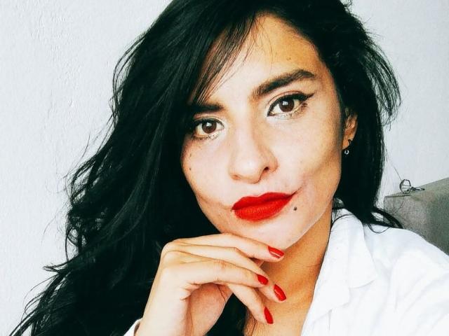 Moserrat Rouse heeft donker, golvend haar, rode lippenstift en rode nagellak, draagt een witte blouse, heeft bruine ogen en een lichte glimlach, waarbij ze haar kin op haar hand rust..