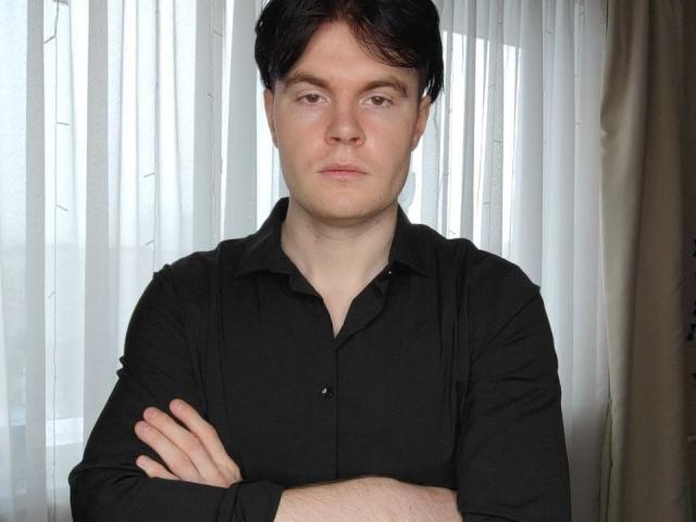 DanielHotS with dark hair, wearing a black button-up shirt, arms crossed, serious expression, standing in front of white vertical blinds.