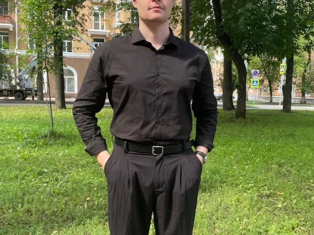 DanielHotS stands in a grassy area wearing a black button-up shirt and pants with hands in pockets. He has a black belt with a silver buckle.