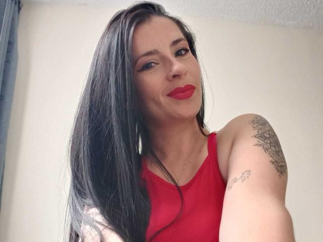 CanellaDiaz with long black hair, red lipstick, and red tank top, showing off tattoos on her arm and shoulder, smiling at the camera.