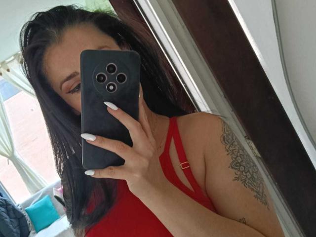 CanellaDiaz with long black hair, wearing a red strappy top, holding a black phone covering her face, showing white nails, and a detailed tattoo on her right arm.