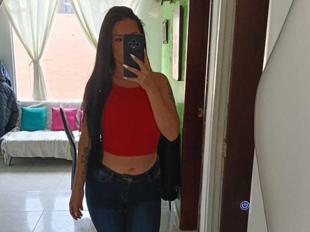 CanellaDiaz with long dark hair, red crop top, and blue jeans, taking a mirror selfie, showing her ass and tattoo on her left arm, holding a black phone.