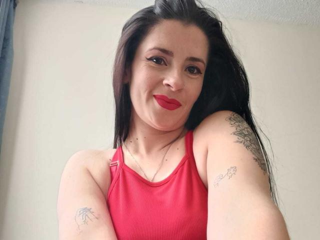 CanellaDiaz with long black hair, red lipstick, and a red tank top, showing tattoos on her arms, smiling at the camera, wearing a necklace.