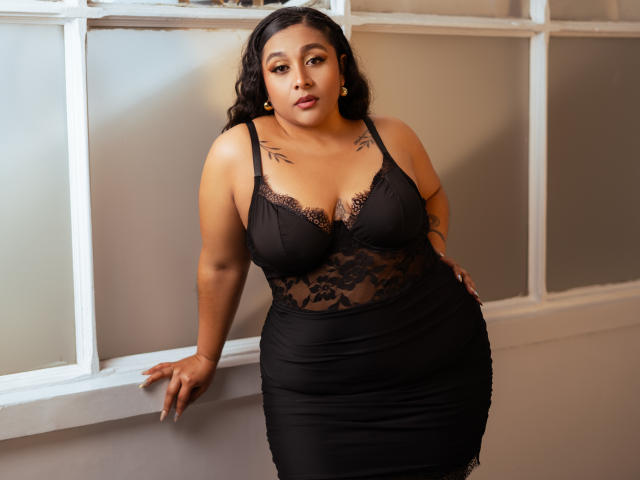 ViickyLee with medium brown skin and curly black hair, wearing a black lace lingerie dress, showing her tits and curvy ass, leaning against a white window frame.