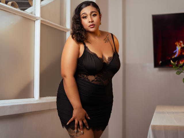 ViickyLee with curvy, brown-skinned body, wearing black lace lingerie, showing her tits and tattoo on left shoulder, standing in room, looking at camera, left hand on thigh.
