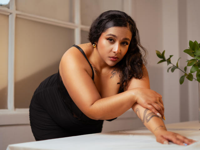 ViickyLee with medium brown skin and black hair, leaning on a table in a black dress, showing her tattoos, with a serious expression and her tits slightly visible.