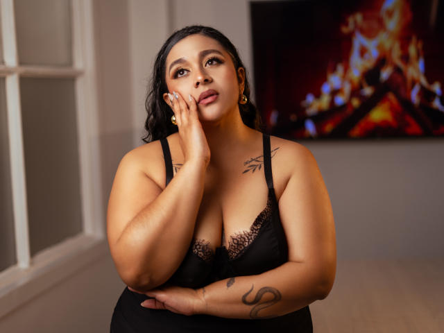 ViickyLee, a curvy woman with medium brown skin and black lace lingerie, touches her cheek with her right hand, showing tattoos on her arm and chest, looking up.