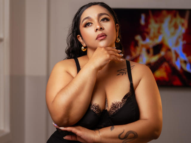 ViickyLee with medium brown skin, black lace bra, large tits, black hair, gold earrings, hand on chin, tattoo on arm, tattoo on chest, looking at camera, confident expression.