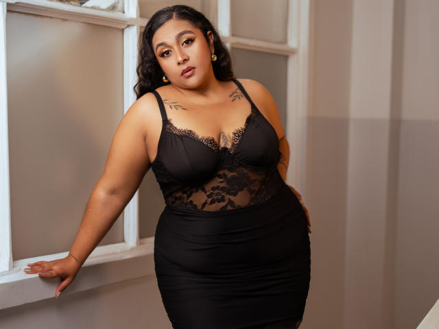 ViickyLee with curvy, tan skin, black lace bustier showing big, round tits, and tight black skirt, leaning against white window, wearing yellow earrings, looking seductive.