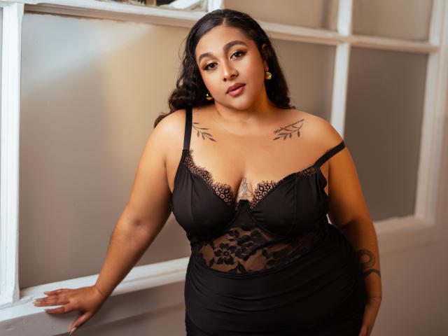 ViickyLee with large tits in black lace lingerie, tattoos on shoulders, dark curly hair, gold earrings, standing against white window frames, looking at camera with a confident expression.