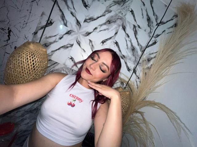 JohanneBlack with pink hair, eyes closed, wearing a white crop top showing small tits, hand on neck, lying down, wicker baskets, and pampas grass in the background.