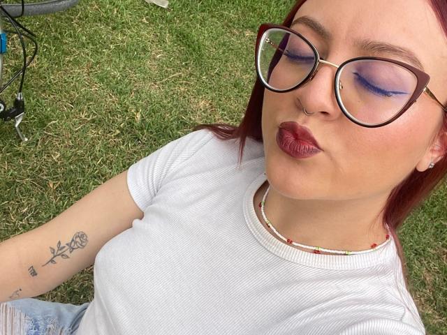 JohanneBlack with purple eyeshadow, pouty red lips, white ribbed shirt, red hair, glasses, nose ring, floral tattoo on arm, sitting on grass, wearing a bead necklace.