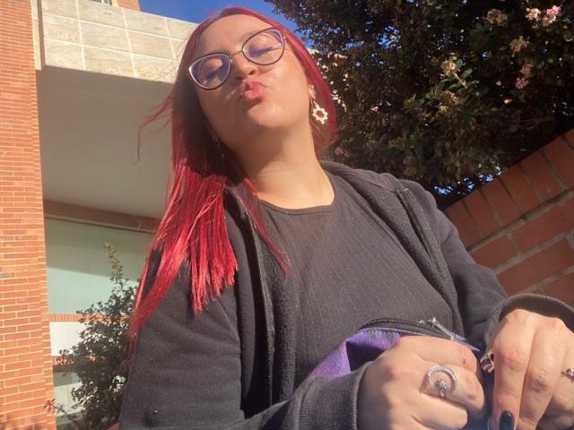 JohanneBlack with red hair, black sweater, and glasses, pouting lips, wearing flower earrings, black jacket, and purple bag, sunlit, holding ring, standing near brick wall.