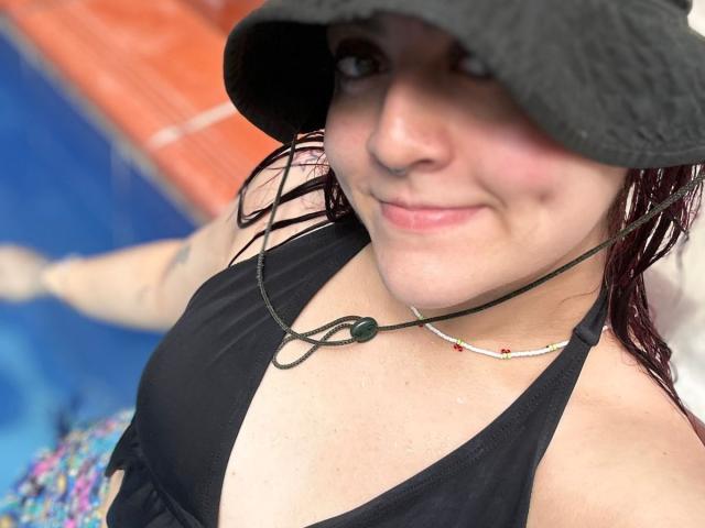 JohanneBlack in a black halter top, wet red hair, black bucket hat, necklace, smiling, showing small boobs, blue pool in background, slightly blurry, light skin, casual look.