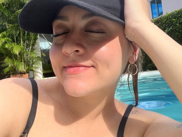 JohanneBlack, with eyes closed, smiling, wearing a black cap, nose ring, and hoop earrings, has wet skin and is by a blue pool, with water droplets on her tits.