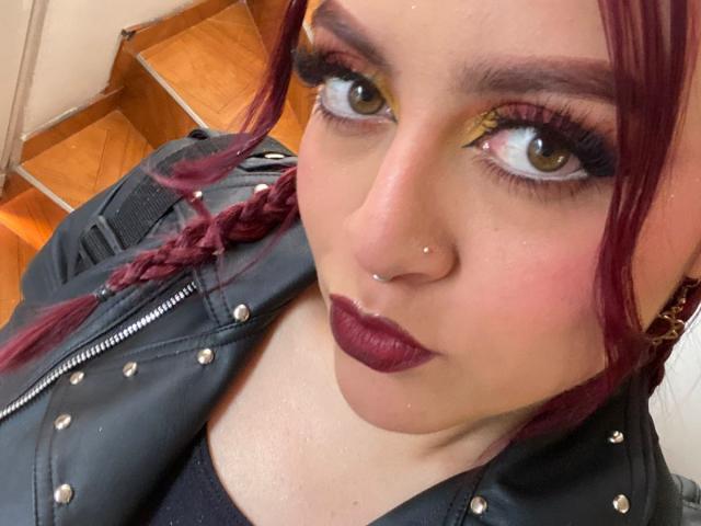 JohanneBlack has red braided hair, dark lipstick, and gold eye makeup. She's wearing a black studded jacket, with a nose piercing and a nose ring. Her eyes are brown.