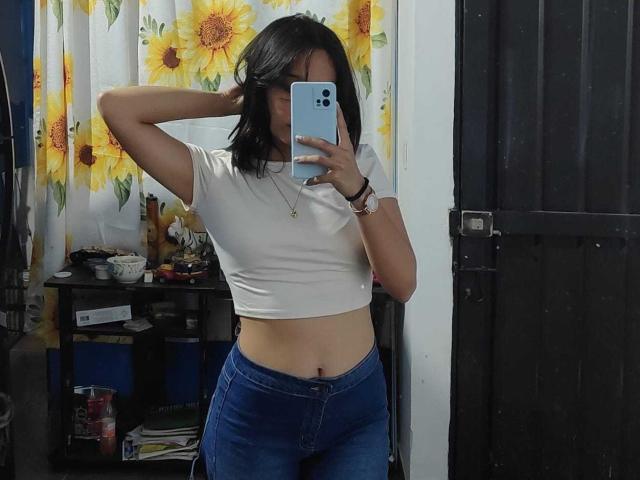 LanaKlaire met zwart haar, een witte crop top, blauwe jeans, die een telefoon vasthoudt en in de spiegel kijkt, waarbij haar navel zichtbaar is, met een gordijn met zonnebloemen op de achtergrond, en haar hand in haar haar..