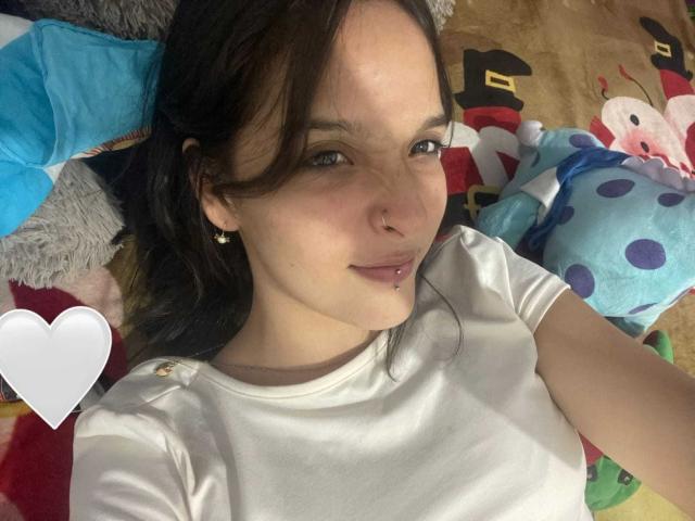 LanaKlaire with pale skin, dark hair, and a nose ring, wearing a white shirt, lying on a colorful blanket, smiling at the camera with a lip piercing.