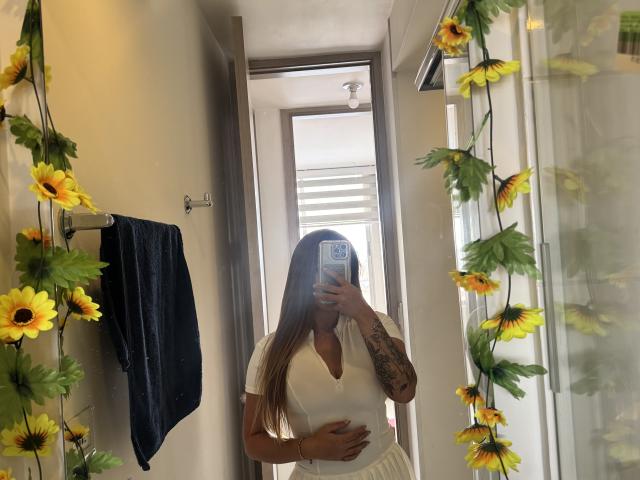 NatSorian with long blonde hair, white V-neck top, showing pregnant belly, tattooed arm, holding phone, sunflower decorations, mirror selfie, blue towel on left.