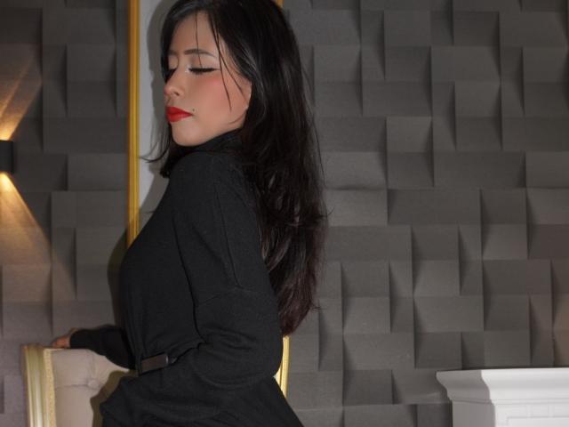CaterinaRomano with long black hair, red lipstick, and black blazer, standing sideways, showing her ass, eyes closed, hand on golden frame, gray textured wall in background.