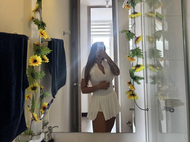 NatSorian with long brown hair, tattooed arm, white dress, showing her tits, holding phone, standing in bathroom with sunflower decor, mirror reflection, hand on belly.