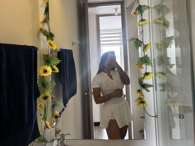 NatSorian, avec sa peau bronzée, son bras tatoué et sa robe blanche, montrant sa poitrine et tenant un téléphone, se tient dans une salle de bain décorée de tournesols et avec une serviette bleue..