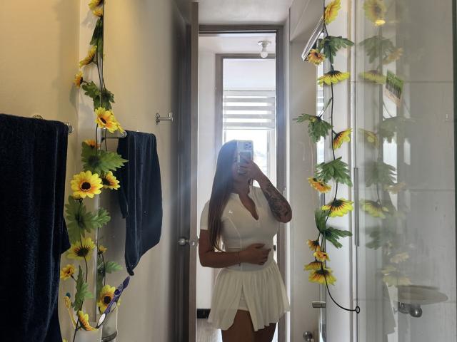 NatSorian con lunghi capelli castani, vestito bianco, che tiene un telefono, coprendosi il seno, specchio decorativo con girasoli, tatuaggio sul braccio destro, asciugamano blu sul lato sinistro, in piedi nel bagno..