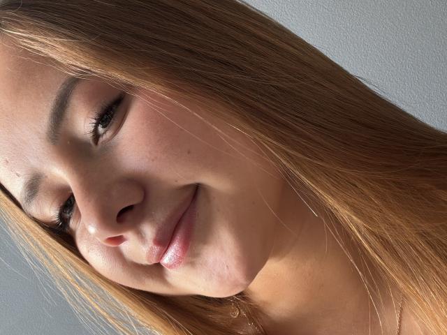 Il viso di NatSorian, con labbra rosate, occhi marroni e lunghi capelli castani, mostra un leggero sorriso, una pelle morbida e un aspetto naturale e delicato..